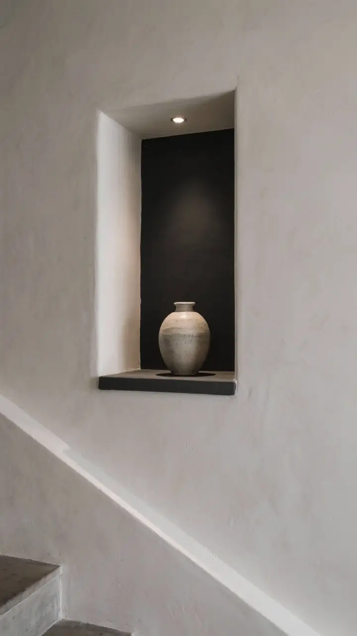 Closeup detail shot of an architectural niche on a landing: interior of the niche painted a contrasting deep charcoal to spotlight a single ceramic vase on a floating ledge. A tiny recessed puck light at the top creates a soft up-lit glow, adding depth and shadow play. Minimalist composition with smooth plaster texture visible.