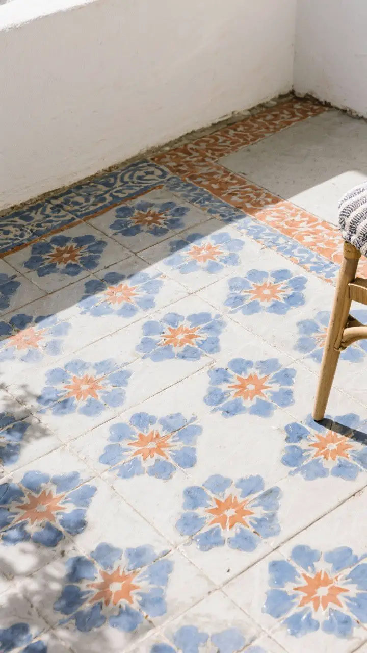 Overhead detail shot of a patterned patio floor: encaustic-look cement tiles with a starburst/floral motif in a cobalt and terracotta palette, slightly matte; a corner shows a budget-friendly stenciled area sealed for durability; nearby textile edge in matching colors to show coordination; bright, even daylight to highlight pattern clarity; no furniture except a hint of chair leg for scale.