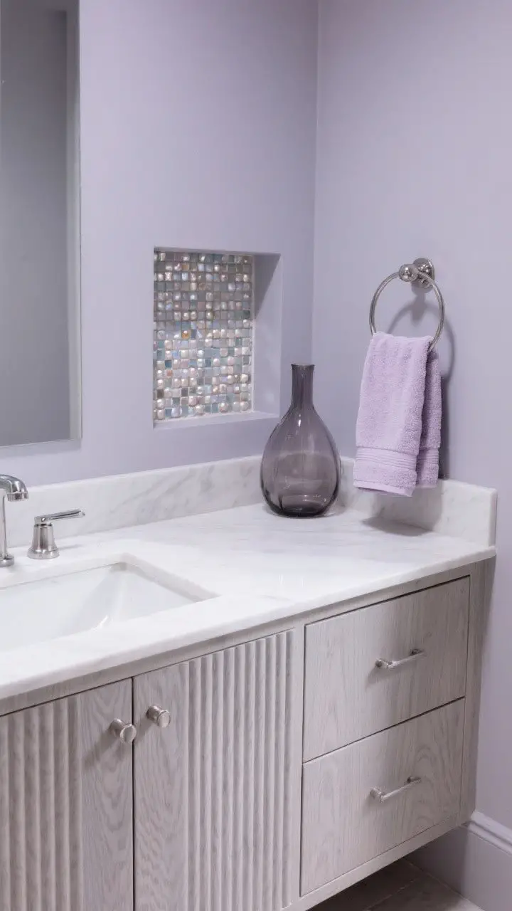Overhead detail shot of a vanity with fluted wood fronts against walls in subtle lavender gray, a niche lined with pearlized mosaic tile shimmering gently, satin nickel pulls, and a smoked glass vase next to ultra-plush pale lilac towels; cool, softly diffused lighting.